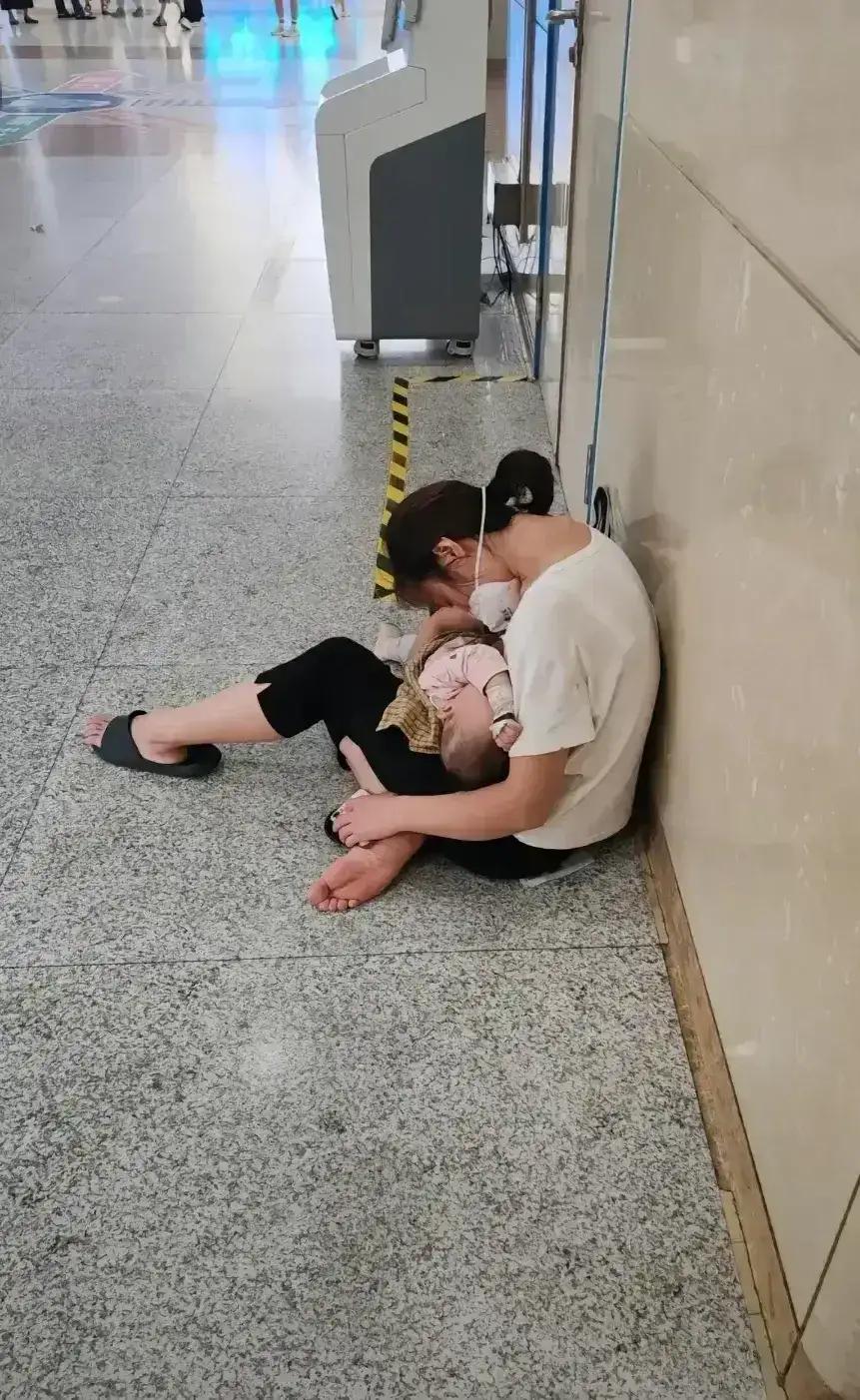 在某医院的一个角落，孩子在妈妈怀抱里安睡，妈妈则坐在地板上。无数来医院就诊的人从