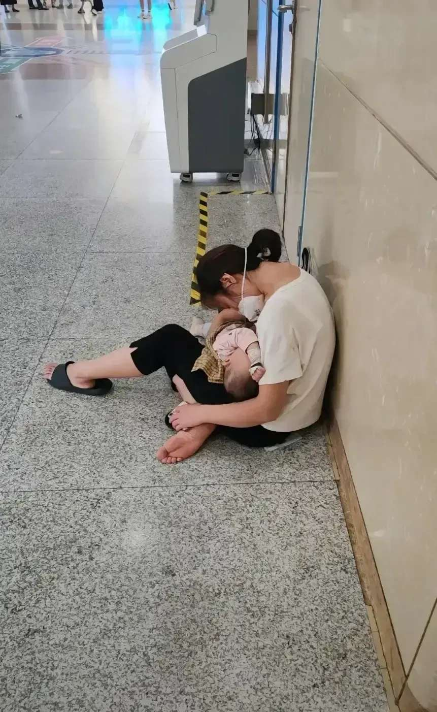 在某医院的一个角落，孩子在妈妈怀抱里安睡，妈妈则坐在地板上。 无数来医院就诊的人