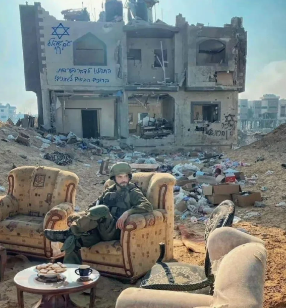 以色列大兵坐在巴勒斯坦人的沙发上晒着太阳喝着下午茶吃着点心，背景的废墟上刷上了以