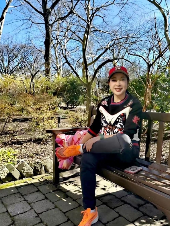 73岁的刘晓庆坐在公园的长椅上留影。镜头下的她身穿黑色上衣搭配黑色裤子，脸上穿着