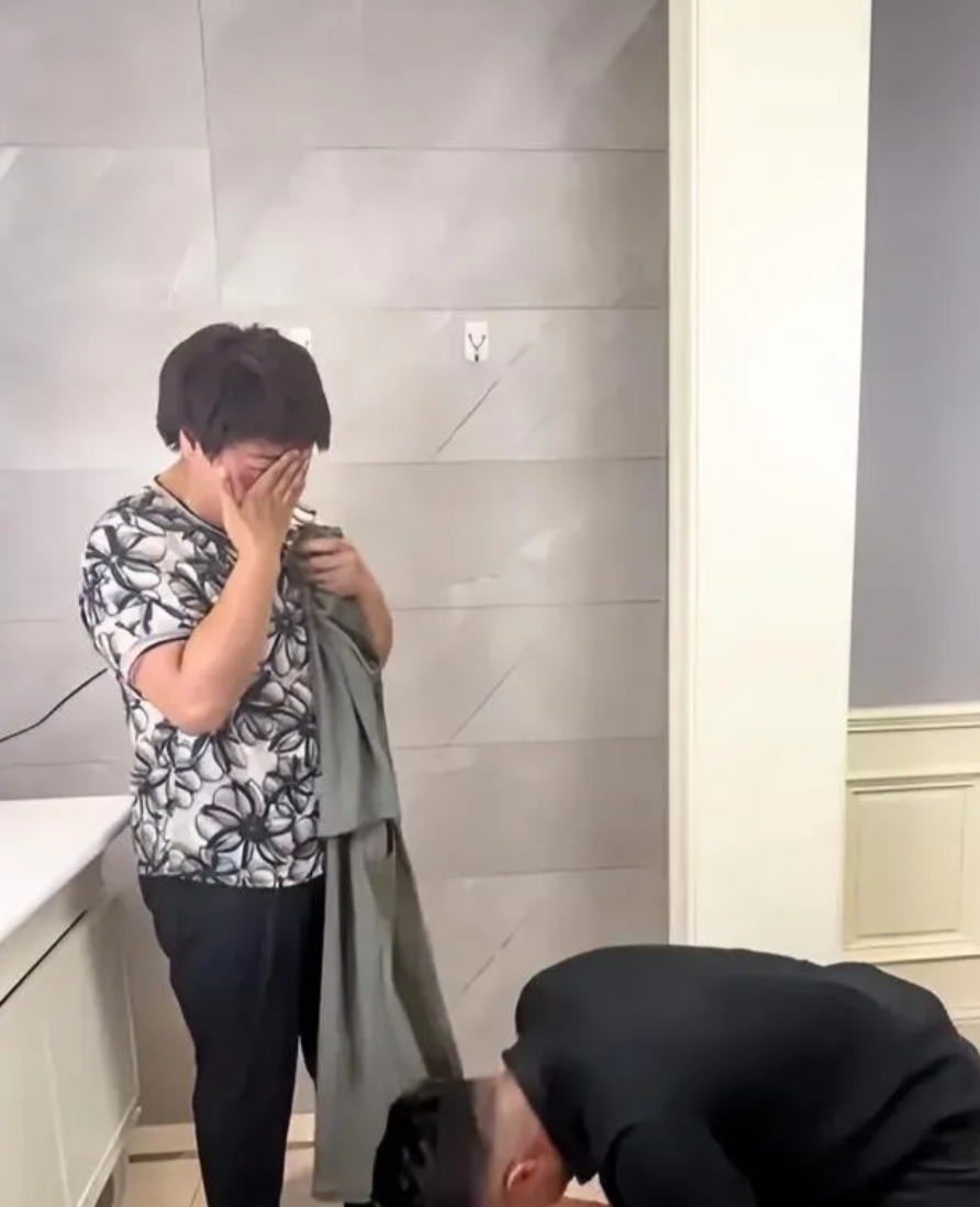 中年女子正在厨房忙碌着，一名年轻男子拿着一件衣服进去后，喊了女子一声，一下就跪倒