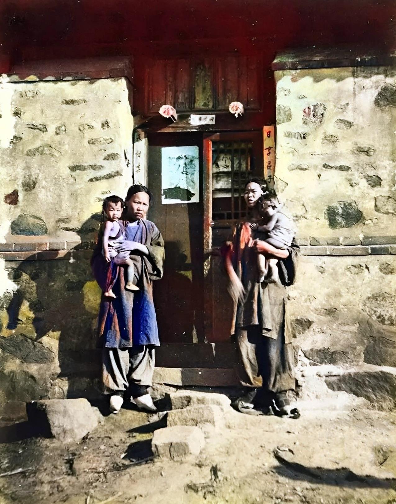 民国，山东农村两名小脚家庭妇女抱着孩子站在堂屋门的两边。堂屋外墙石头外露，门窗破