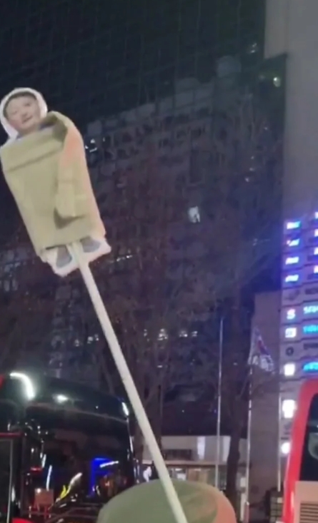 在一次首尔抗议中，有市民举着被草席卷着的尹锡悦的人型牌子。

尹大统领身穿囚服，