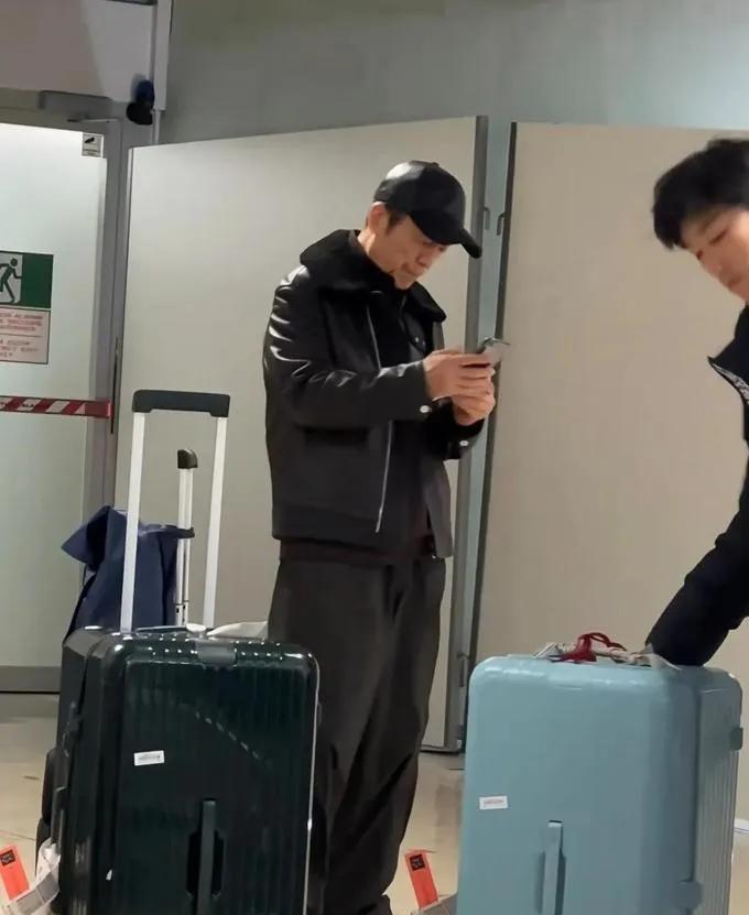 知名导演张艺谋现身日内瓦机场被网友偶遇，头戴黑色棒球帽，黑色皮衣外套，手里拿着手