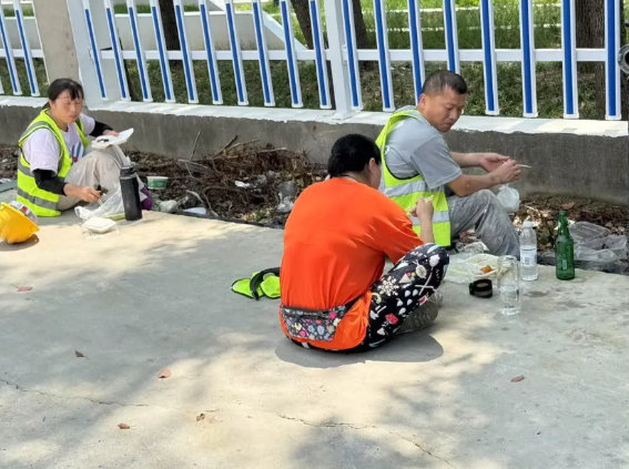 工地中午吃饭时间，工人坐在马路边吃着盒饭喝着啤酒，正好被路过的业主看到了，随即询