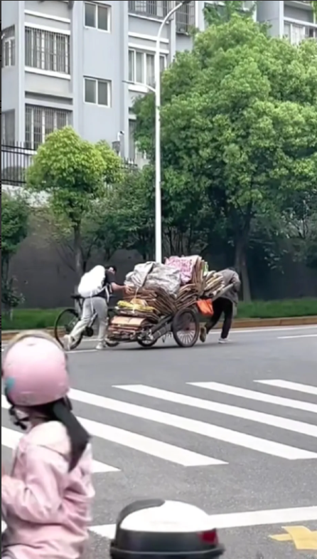 上坡路段，白发老人佝偻着腰，拉着一车废纸皮，显得有些吃力了，路过学生娃一个举动，