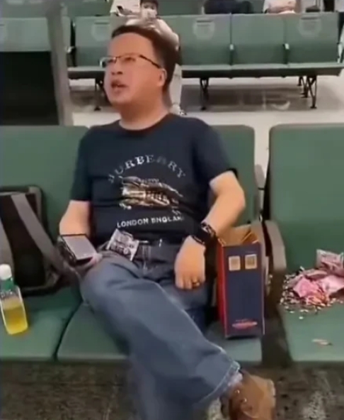 深圳宝安机场候机大厅内，一位戴着眼镜穿着考究的男子坐在椅子上看手机，旁边的座位上