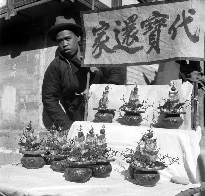 民国，一名小摊贩摆摊卖聚宝盆。他还打出了“代宝还家”的横幅。小贩并没有写“买宝还