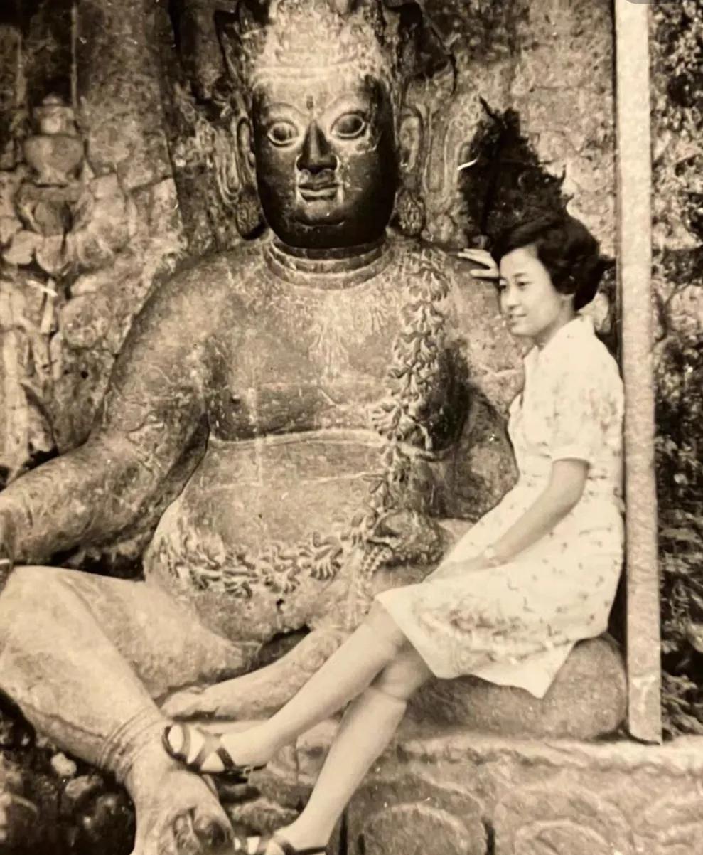 面容姣好的年轻女子坐在大佛单盘的左腿上。她面带微笑，身着素雅的白色连衣裙，下身搭