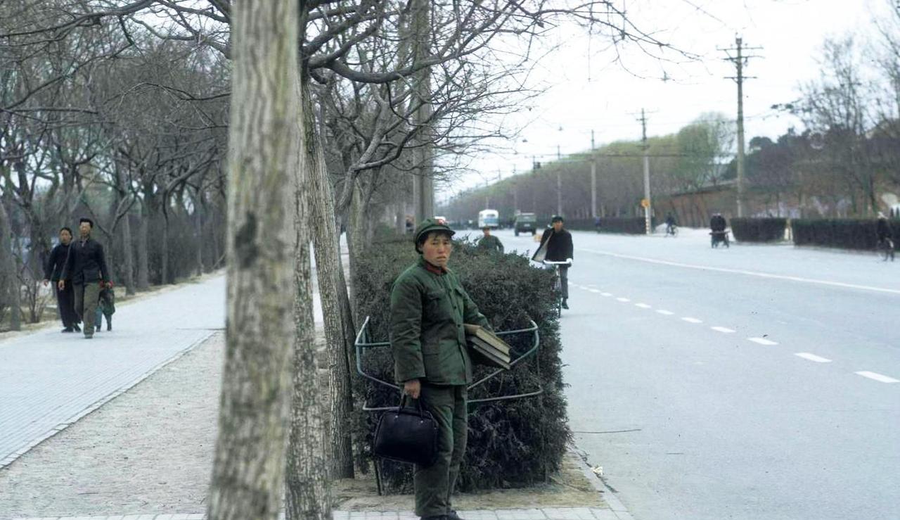1972年，北京街头，一名女子在路边等公交车。她身穿65式军服，右手拎黑色提包，