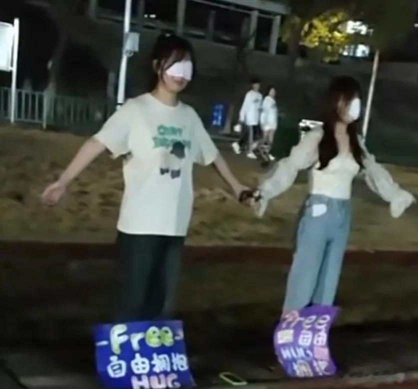 某校操场上，两名女生手拉手，呈张开姿态；两人脚边各放着一个牌子，上面写着“自由拥