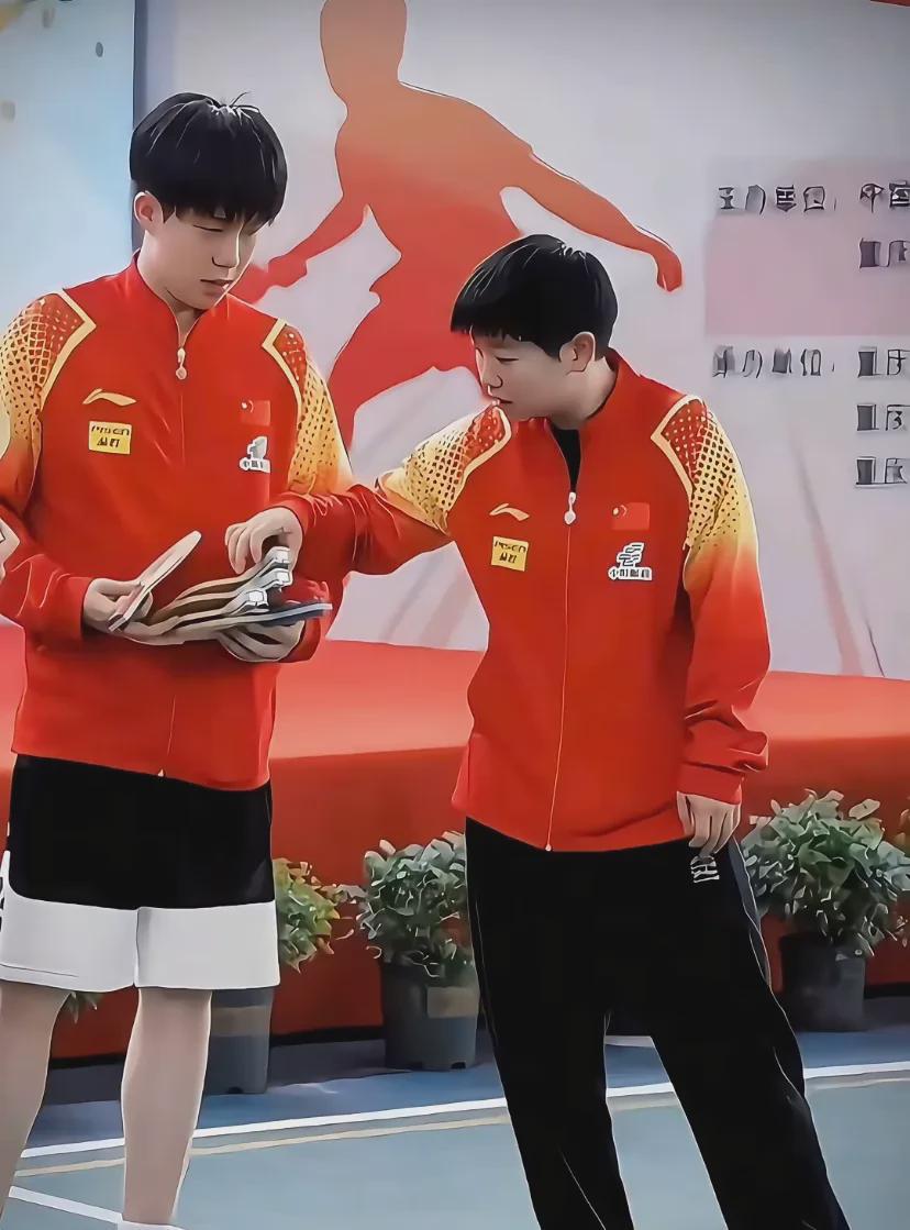 小萨在王楚钦衣兜里取零食，手中握着球拍，王楚钦无动于衷，只是旁观。
她取，他看