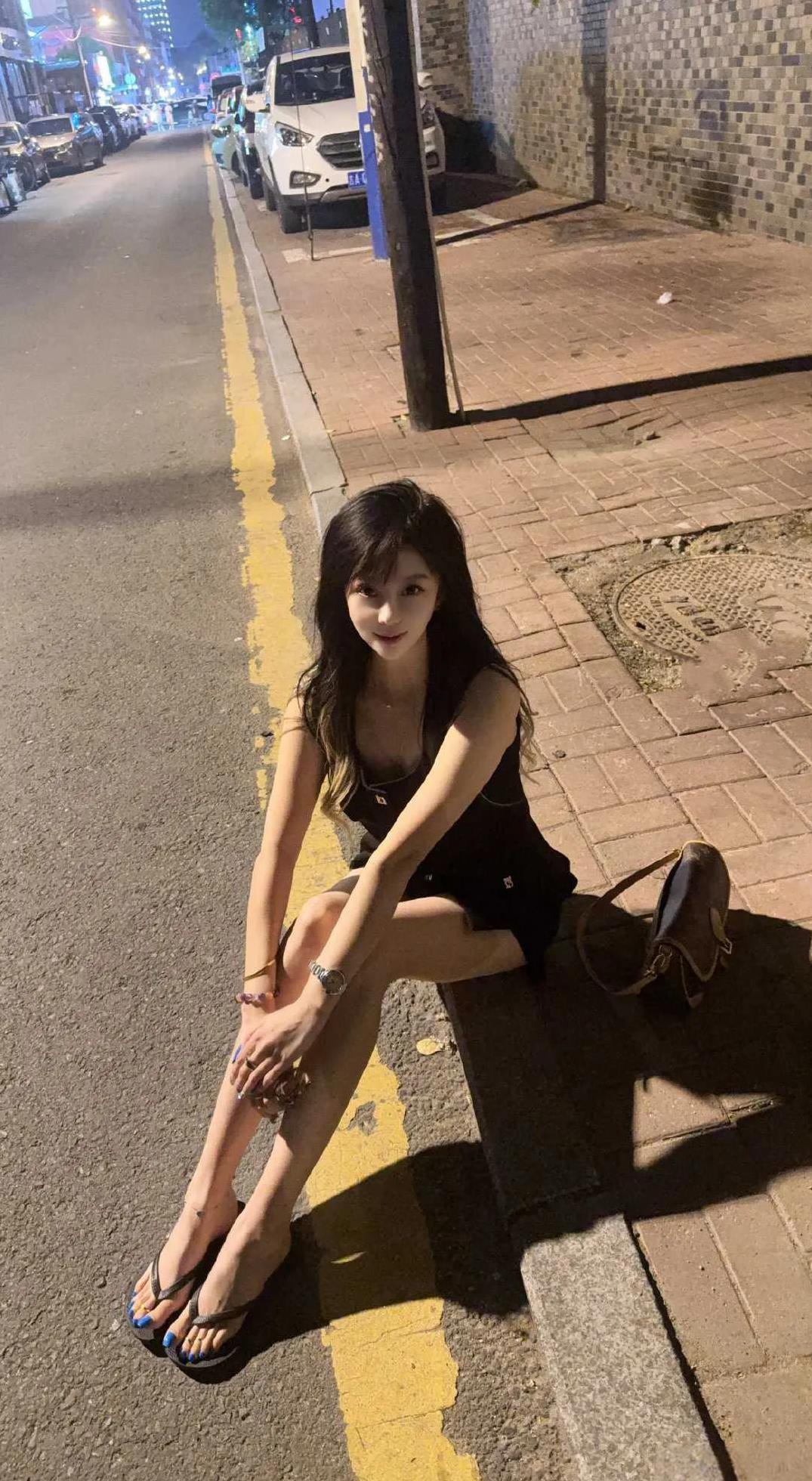 夜色街头，黑衣小姐姐静坐路边，发丝微卷，笑意藏在眉眼。城市喧嚣成背景，她自成静谧