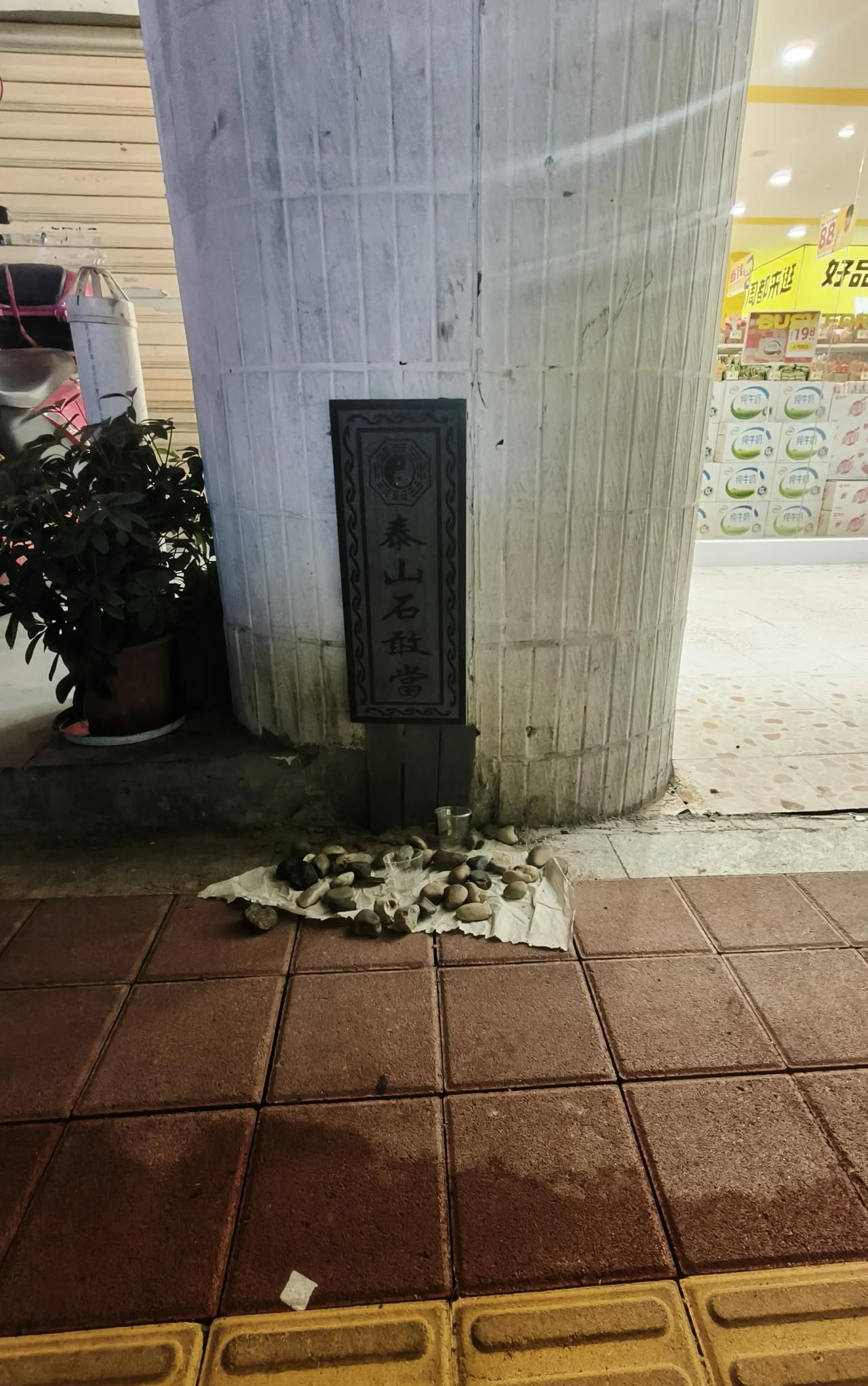 某栋大楼柱子前摆放的物件:一堆石子、两只塑料杯，后背“泰山石敢当”五个大字上配有