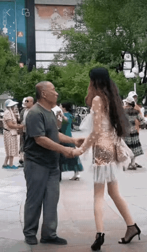 公园里面，有个老头和一名身材纤细、穿着时髦的女子正在跳广场舞。老头紧紧握着女子的