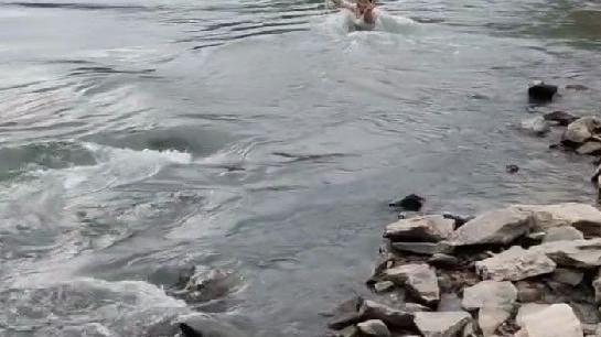 女子跳入水流湍急的河中，被急流的河水冲到下游，还朝岸上的人笑