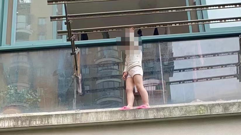 小女孩站在自家阳台外，双手紧紧握住阳台的晾衣架……