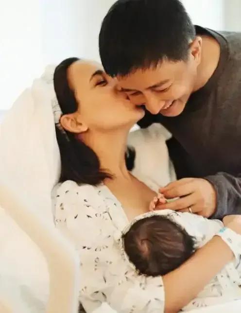 |妈妈卢靖姗则一边抱着宝宝一边亲吻老公脸颊，画面幸福温馨十足