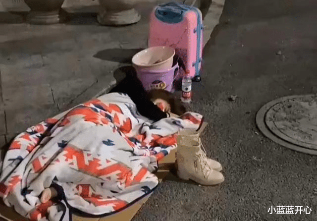 年轻女子露宿街头,身下垫着纸板,身旁放着生活用品和行李箱