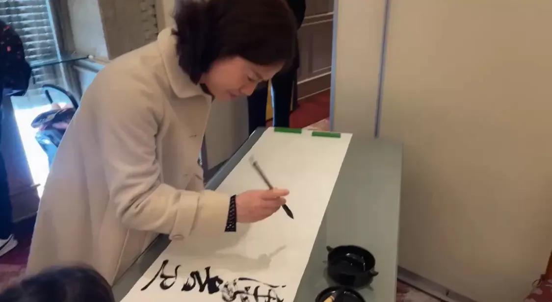 翁帆在香港参加春茗，她穿白色大衣很美，她写完毛笔字害羞的笑了