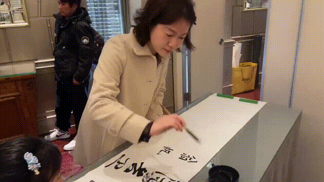 翁帆在香港参加春茗，她穿白色大衣很美，她写完毛笔字害羞的笑了